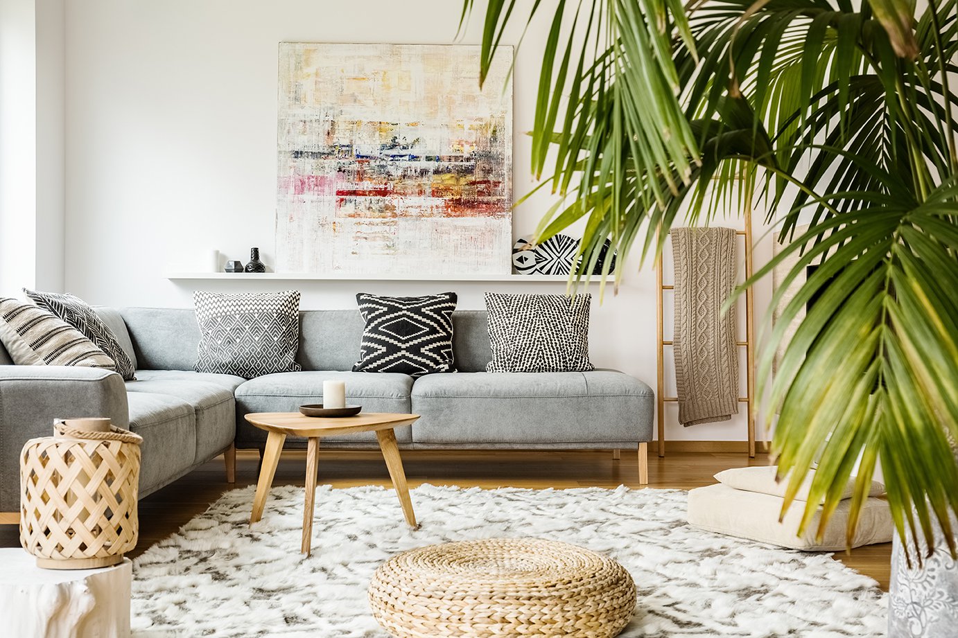 Pouf and wooden table in modern living room with painting above grey corner couch.